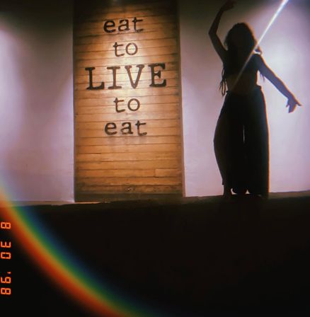 Dimly lit cafe wall with wooden sign reading "eat to LIVE to eat" and a silhouetted dancer posed beside it, warm backlighting and a rainbow lens flare