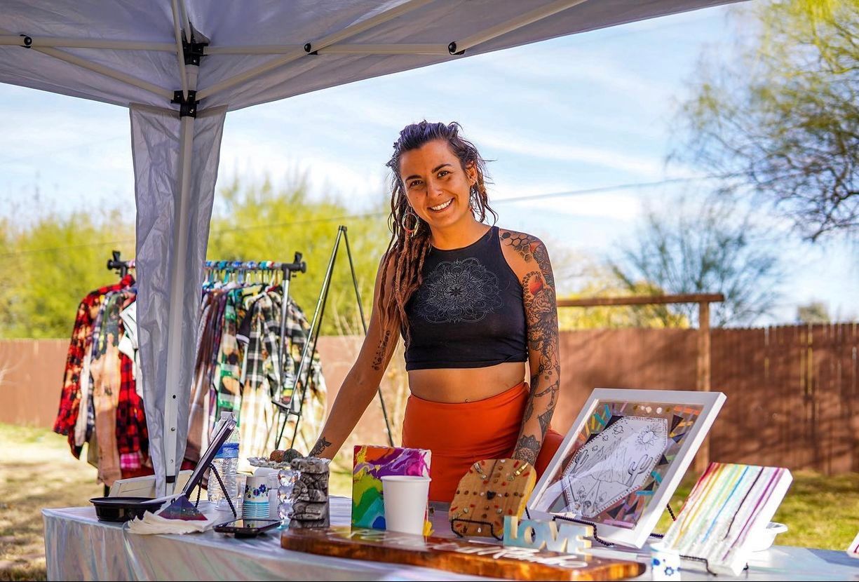 Smiling vendor with dreadlocks and arm tattoos stands behind a colorful outdoor craft market booth under a canopy, displaying handmade art prints, jewelry and clothing racks on a sunny day.