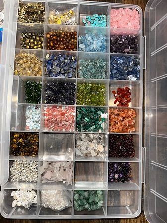 Clear plastic compartment organizer filled with colorful gemstone and crystal beads — polished chips, pearls and round beads neatly sorted for jewelry making and craft projects in a craft studio.