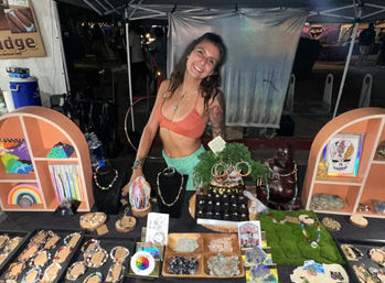 Smiling vendor at a night market jewelry booth displaying handmade beaded necklaces, rings, bracelets, gemstone clusters and colorful art under a canopy