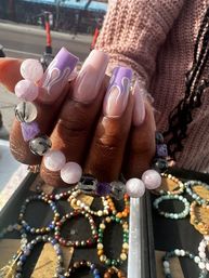 Hand with long nude-to-lavender coffin acrylic nails with white flame tips holding pink and gray beaded gemstone bracelets over a tray of colorful beaded bracelets at an outdoor market stall