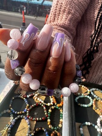 Hand with long nude-to-lavender coffin acrylic nails with white flame tips holding pink and gray beaded gemstone bracelets over a tray of colorful beaded bracelets at an outdoor market stall