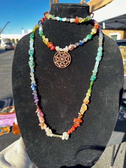 Rainbow gemstone chip necklaces with a wooden mandala pendant displayed on a black velvet bust at an outdoor market under a bright blue sky