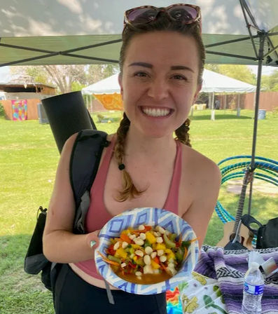 Smiling woman at a sunny outdoor park market holding a colorful corn-and-bean salad in a paper bowl under a canopy, with hula hoops, guitar and tents in the background.