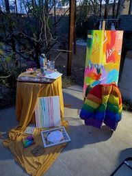 Backyard patio art display with bright abstract canvases, paint supplies on a yellow-draped table, a tall colorful painting on an easel, and a rainbow puffer jacket draped over a chair.