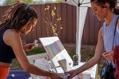 Sunlit outdoor craft market stall under a canopy as a vendor hands a small bowl to a paying customer, colorful art prints displayed on an easel