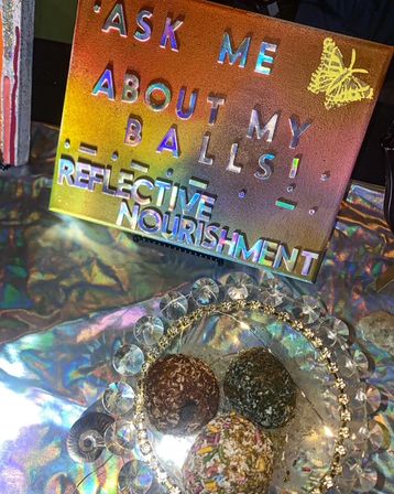 Holographic sign reading Ask me about my balls and Reflective Nourishment above a crystal platter of three sprinkle- and coconut-coated energy balls on an iridescent vendor table.