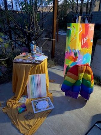Backyard patio art display with bright abstract canvases, paint supplies on a yellow-draped table, a tall colorful painting on an easel, and a rainbow puffer jacket draped over a chair.