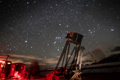 Dobsonian telescope on a tripod at a dark beach campsite with red gear lights and blurred figures, framed by a star-filled night sky for coastal stargazing.