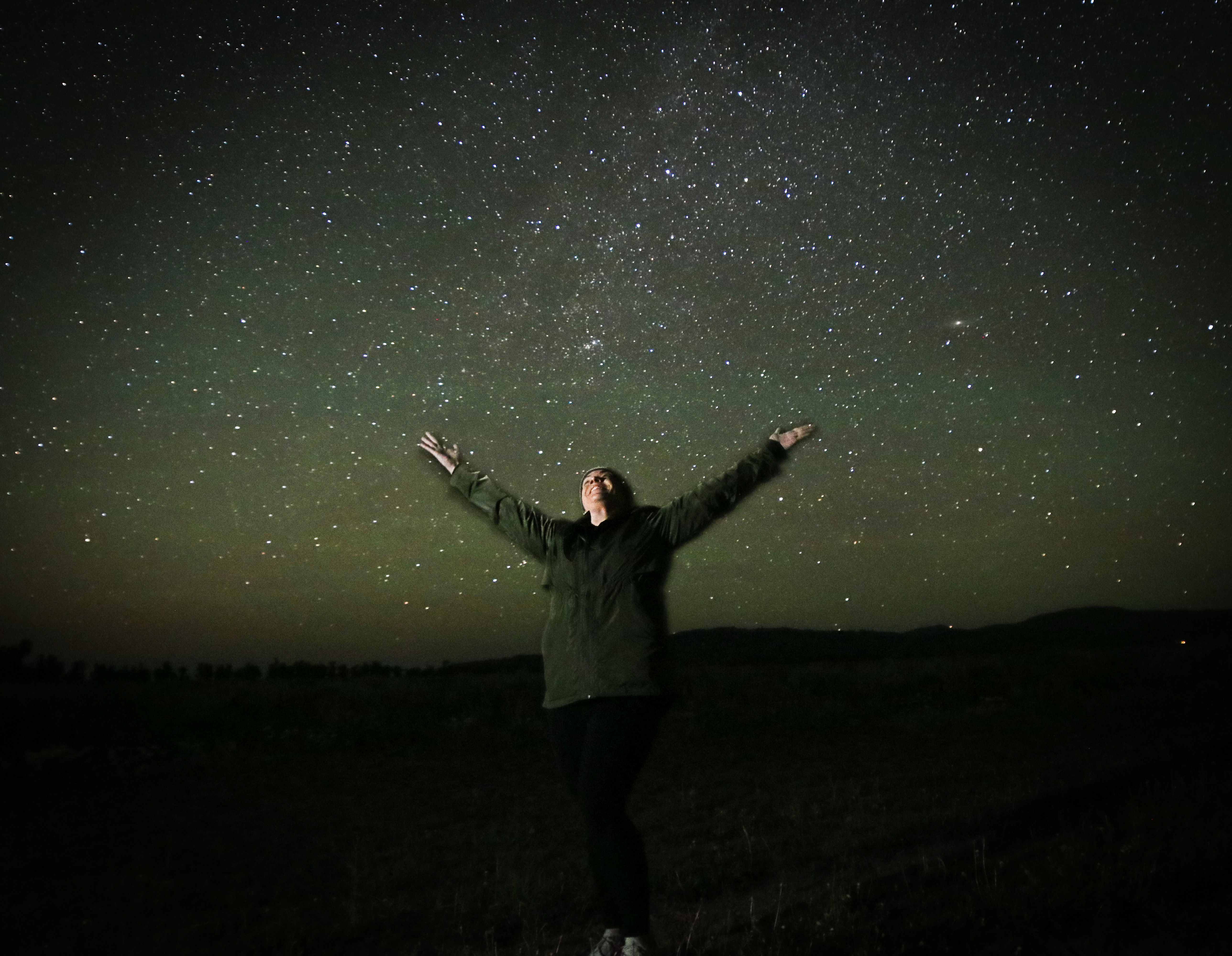 Person standing in a dark rural field with arms outstretched, looking up at a vibrant star-filled night sky with the Milky Way visible.