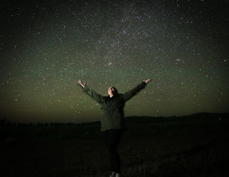 Person standing in a dark rural field with arms outstretched, looking up at a vibrant star-filled night sky with the Milky Way visible.