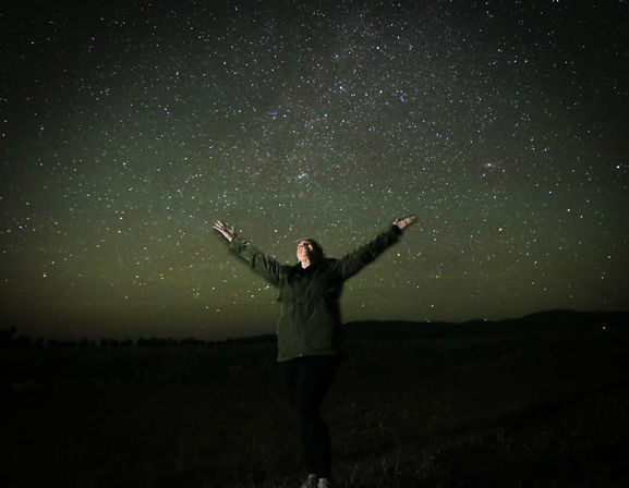 Person standing in a dark rural field with arms outstretched, looking up at a vibrant star-filled night sky with the Milky Way visible.