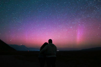 Silhouetted couple embracing under pink and purple aurora borealis with a starry night sky and dark mountain horizon