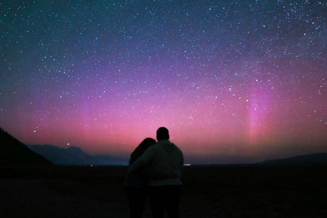 Silhouetted couple embracing under pink and purple aurora borealis with a starry night sky and dark mountain horizon