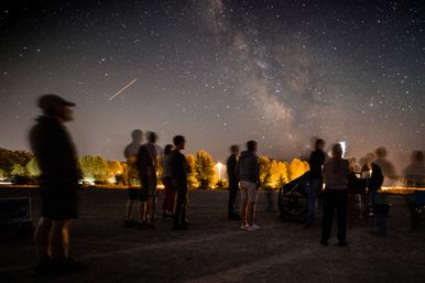 Outdoor stargazing event under the Milky Way — long-exposure blur of spectators around a telescope, illuminated treeline and a bright shooting-star streak in the night sky