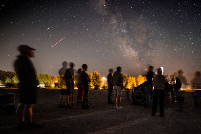Outdoor stargazing event under the Milky Way — long-exposure blur of spectators around a telescope, illuminated treeline and a bright shooting-star streak in the night sky