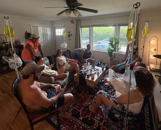 Group of friends lounging in a sunny suburban living room around a coffee table, some seated with IV drip bags on stands, chatting on couches and chairs with a potted plant and parked cars visible outside the window.