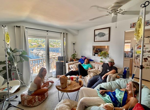 Cozy lakeside living room with four friends lounging on sofas and floor, IV drip stands beside them, round wooden coffee table, sliding doors opening to a marina view