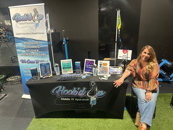 Wellness booth at an indoor gym health fair featuring an IV hydration setup with a hanging IV bag, brochures and wellness products on a black tablecloth, and a smiling attendant seated beside the display.