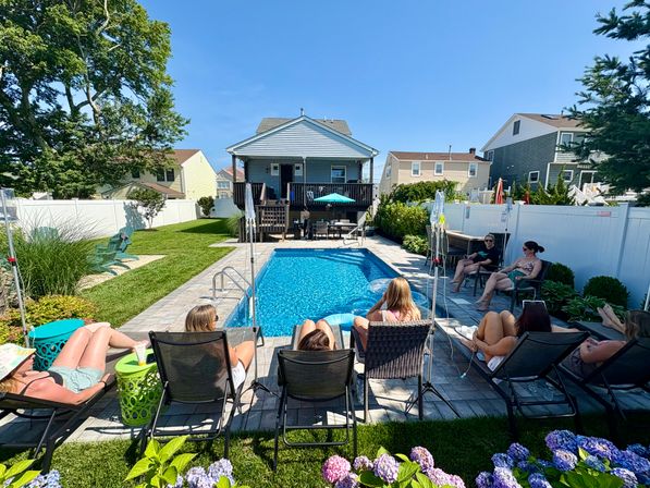 Sunny suburban backyard with rectangular in-ground pool, people lounging on deck chairs, two-story house with deck and umbrella, white privacy fence, green lawn and hydrangea blooms.