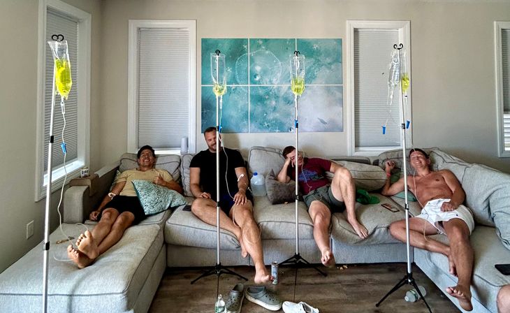 Four friends lounging on a gray sectional sofa in a bright living room receiving yellow vitamin IV drips on poles, feet up and laughing with shoes and water bottles on the floor