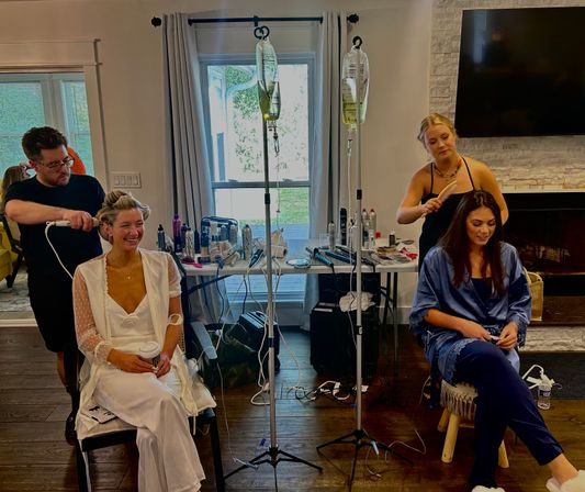 Wedding morning at-home prep: person in a white robe laughing with coffee while a stylist curls her hair, another person in a blue robe has hair brushed, IV hydration bags on stands and hair tools spread on a table in a cozy living room.
