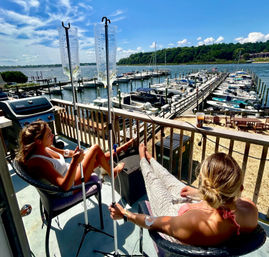 Two women lounging with IV drip bags on a sunny waterfront balcony, feet up and relaxing while overlooking a busy marina with boats and wooden docks under a blue sky