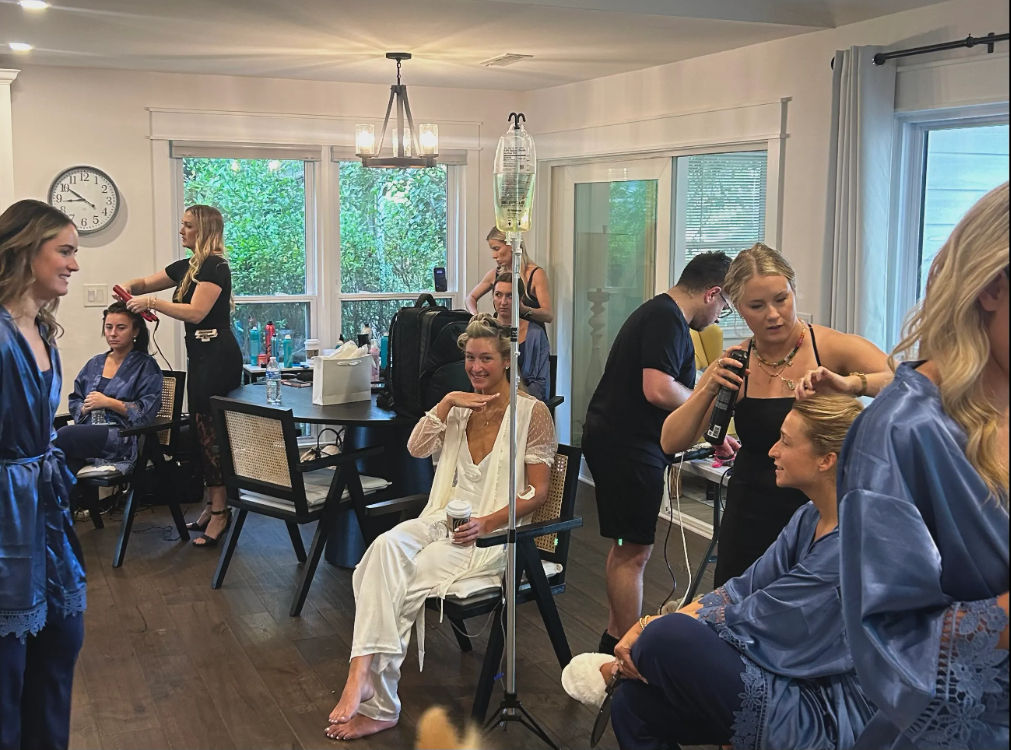 Wedding morning bridal party in a bright living room — women in robes having hair and makeup done, bride in white sipping coffee beside an IV drip stand as stylists work.
