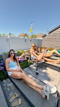 Four women in swimsuits lounging on poolside chaise lounges in a sunny suburban backyard with IV hydration drips on poles, purple hydrangeas, white privacy fence and stone paver patio under a clear blue sky
