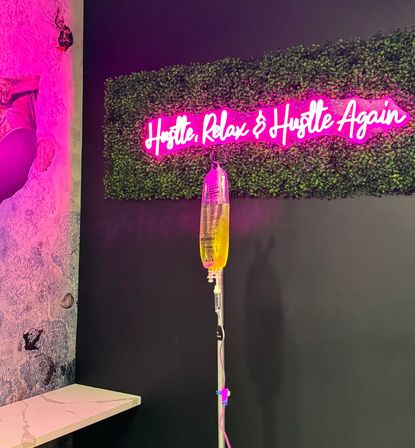 Aesthetic wellness lounge corner with a pink neon sign reading Hustle, Relax & Hustle Again mounted on a faux boxwood wall, pink ambient lighting and a yellow IV drip bag on a stand in the foreground.