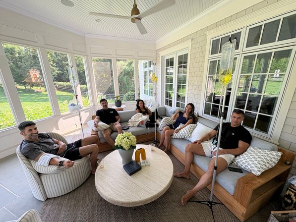 Five adults relaxing and receiving IV drips in a bright screened-in sunroom/porch, seated on a sectional and chairs around a round coffee table with flowers and large windows looking out to a green backyard.