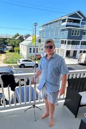 Person in sunglasses and a baseball cap stands barefoot on a sunny beach-town balcony holding a clear IV-style drip bag with yellow fluid on a tripod, parked cars and multi-story coastal houses visible in the background.