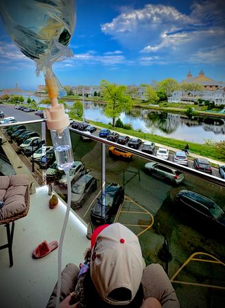 Close-up IV bag and tubing on a balcony with a person in a baseball cap, overlooking a riverfront neighborhood with parked cars, tree-lined shore and bright blue sky.