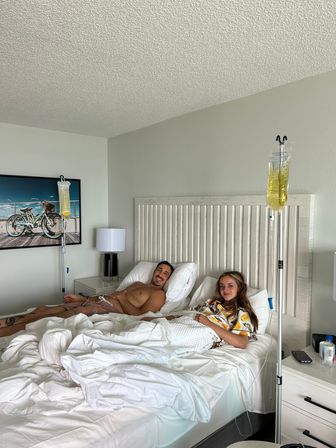 Smiling couple reclining on rumpled white bedding in a bright coastal-style bedroom, each with an IV pole holding yellow IV bags, beach bicycle artwork on the wall.