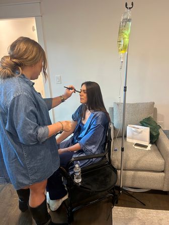 Makeup artist applying eye makeup to a seated woman in a blue robe beside an IV drip on a stand during an at-home beauty session in a cozy living-room setting.