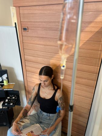 Person with tattooed arms sits in a wooden-paneled wellness lounge reading a magazine while receiving an IV drip, wearing a black tank top and ripped jeans.
