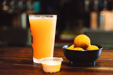 Hazy orange craft beer in a pint glass with a black bowl of golden fried croquettes and a small dipping sauce cup on a wooden bar counter, bar snack scene.
