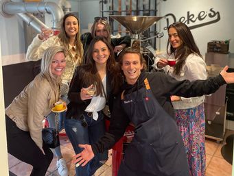 Smiling group of six adults holding espresso cups and drinks inside an artisanal coffee roastery, posing in front of a large coffee roasting machine.