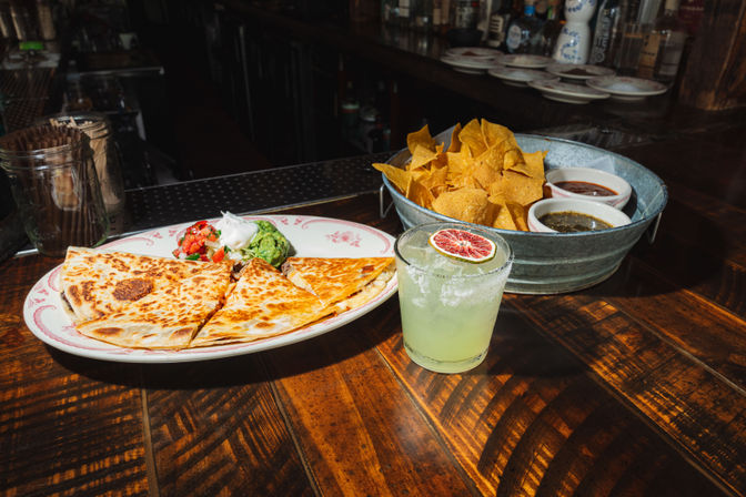 Cheesy quesadilla wedges with guacamole, pico de gallo and sour cream on a plate beside a metal bucket of tortilla chips and two salsas, plus a salted-rim margarita garnished with a dried blood orange slice on a wooden bar counter.