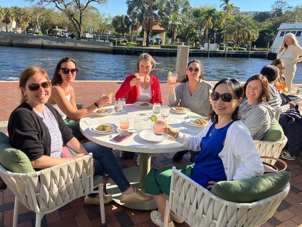 Smiling group of women toasting cocktails at a sunny waterfront patio brunch, plates of food on the table, palm trees and a river in the background.