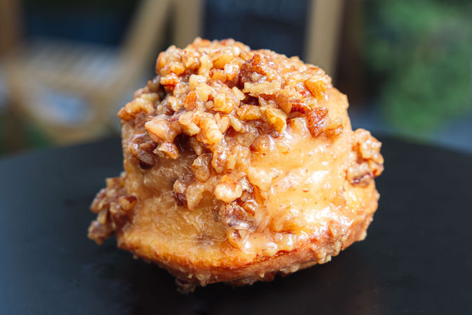 Close-up of a gooey glazed pecan sticky bun topped with chopped pecans on a black plate, bakery-style pastry shot.