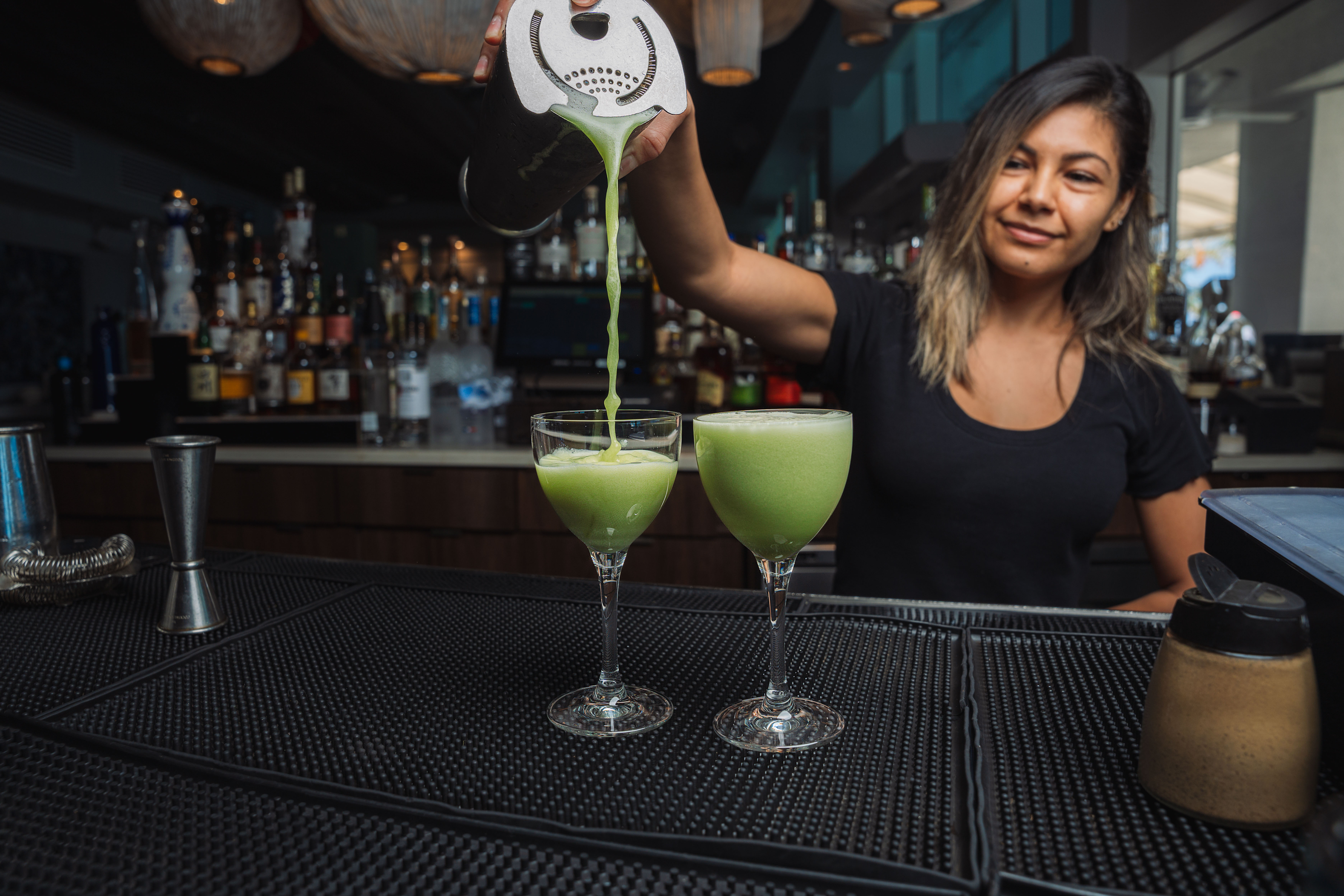 Bartender pouring vibrant green cocktails from a shaker into coupe glasses on a busy bar counter