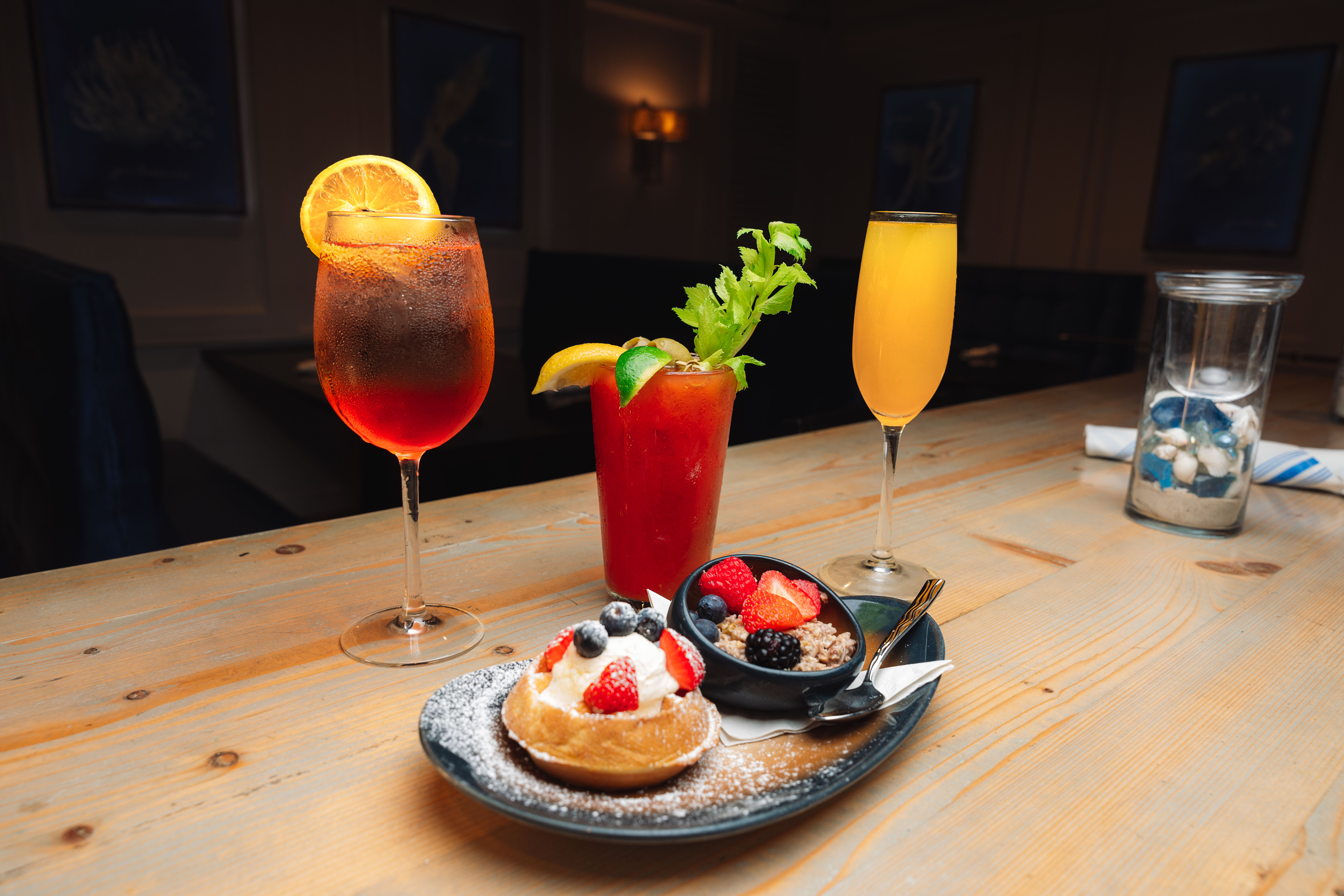 Brunch cocktails on a light wooden bar: chilled orange spritz with an orange slice, Bloody Mary with celery, mimosa flute, and a plate with a cream-topped pastry, fresh berries and granola.