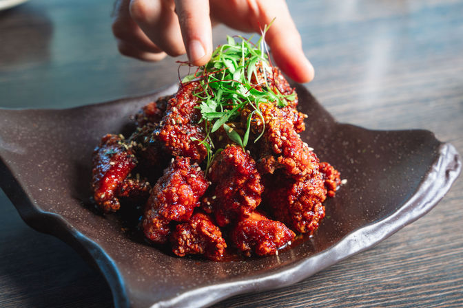 Crispy spicy glazed Korean-style fried chicken piled on a dark ceramic plate, sprinkled with sesame seeds and microgreens, a hand reaching to pick a piece.
