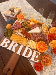 Festive bridal shower charcuterie board on a wooden paddle with assorted cheeses, cured meats, crackers, olives, pickles, pistachios, citrus slices, jam, mini desserts, champagne flutes and orange floral accents.