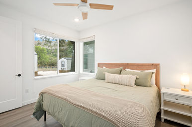 Cozy bright bedroom with wooden headboard, sage-green bedding and chunky knit throw, bedside table with lamp, corner windows overlooking trees and a small outbuilding.