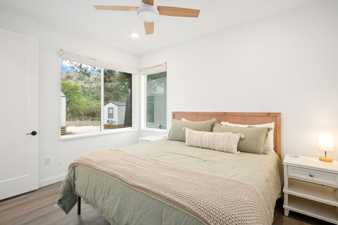 Cozy bright bedroom with wooden headboard, sage-green bedding and chunky knit throw, bedside table with lamp, corner windows overlooking trees and a small outbuilding.