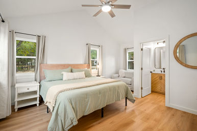 Bright, airy bedroom with sage-green bedding, light wood headboard, ceiling fan, large windows, cozy armchair, warm wood floors and en‑suite bathroom