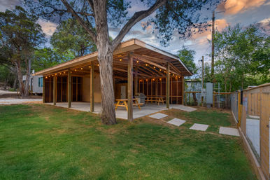 Covered outdoor pavilion with string lights, wooden picnic tables and a grill on a concrete slab in a grassy, tree-lined yard at dusk