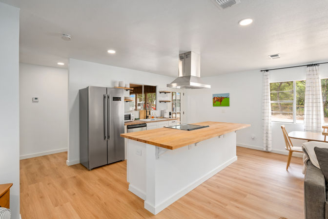 Sunlit open-concept kitchen with a large butcher-block island and cooktop, stainless steel refrigerator, ceiling range hood, light wood floors, white walls, windows with curtains, and a cozy dining nook with colorful cow artwork.